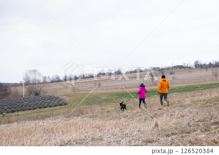 Joyful stroll through a tranquil countryside with a child and a dog on a cloudy day, surrounded by expansive fields and trees in the distance during early spring Joyful stroll through a tranquil countryside with a child and a dog on a cloudy day, surrounded by expansive fields and trees in the distance during early spring 126520384