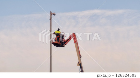 Worker in safety gear operating at height on lift, servicing pole equipment against clear sky backdrop 126524305