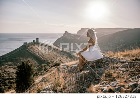 A woman is sitting on a hillside overlooking the ocean. She is wearing a white dress and has blonde hair. The scene is serene and peaceful, with the ocean in the background. 126527818