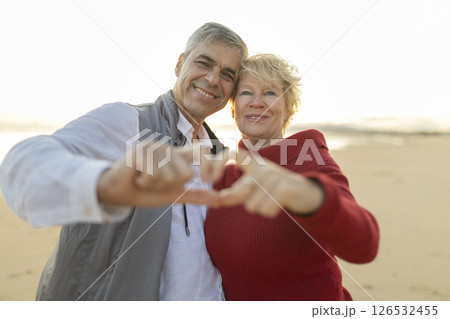 Happy senior couple making heart shape with hands on beach at sunset 126532455