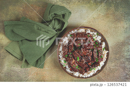 crispy beef, with green onions and sesame seeds, on a rice pad, in a bowl with chopsticks, on a textured surface, no people 126537421