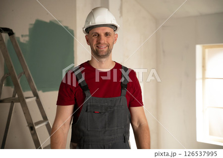 Smiling contractor wearing hardhat and overalls in unfinished room Smiling contractor wearing hardhat and overalls in unfinished room 126537995