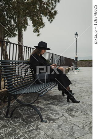 Elegant Woman With A Black Hat Using The Smartphone While Sitting On The Bench  126546025