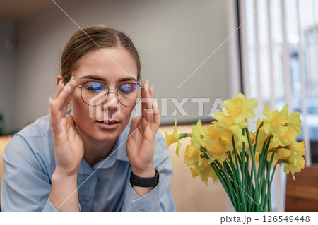 Headache woman glasses: Woman rubs temples indoors, likely due to a headache, near yellow flowers. Headache woman glasses: Woman rubs temples indoors, likely due to a headache, near yellow flowers. 126549448