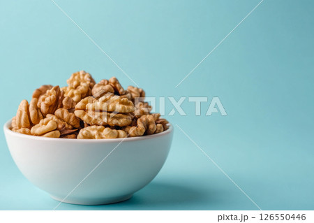A white bowl filled with walnuts sits on a light blue background. A white bowl filled with walnuts sits on a light blue background. 126550446