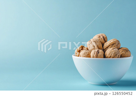 A white bowl filled with walnuts sits on a light blue background. A white bowl filled with walnuts sits on a light blue background. 126550452