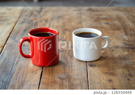 Two ceramic mugs, one red and one white, sit side by side on a wooden table, with a softly blurred background featuring brick walls and greenery. 126550489