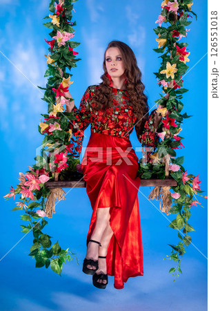 An Elegant Woman Posing Gracefully on a Floral Swing While Wearing a Stunning Red Dress 126551018