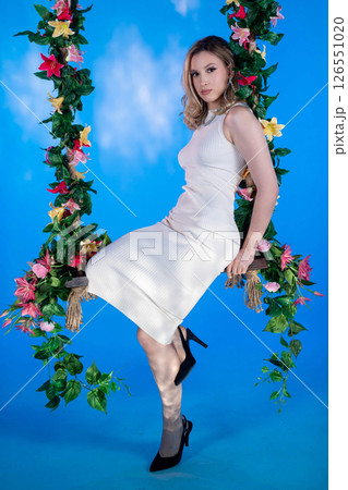 An Elegant Woman Posing Gracefully on a Floral Swing Against a Bright Blue Background 126551020