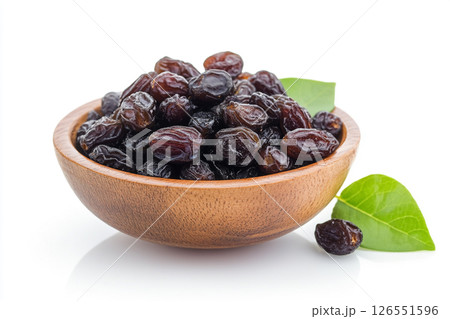 A wooden bowl filled with glossy, dark dates is surrounded by green leaves, with a few dates scattered outside the bowl. 126551596