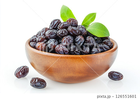 A wooden bowl filled with glossy, dark dates is surrounded by green leaves, with a few dates scattered outside the bowl. 126551601
