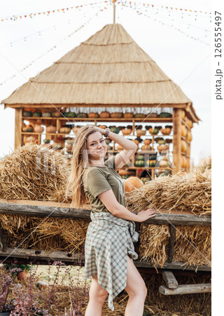 young woman stands at pumpkin festival, holding large orange and green pumpkin. She is surrounded by rows of pumpkins and a wooden display. The autumn farm setting captures the harvest season vibe. young woman stands at pumpkin festival, holding large orange and green pumpkin. She is surrounded by rows of pumpkins and a wooden display. The autumn farm setting captures the harvest season vibe. 126555497