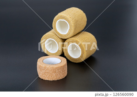 Four elastic medical bandage rolls are stacked on dark surface, showcasing their texture and cylindrical shape. These bandages are commonly used for support and compression in medical settings 126560090