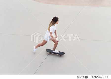 Skateboarder girl in a white t-shirt and denim shorts rides a skateboard in modern urban skatepark. 126562254