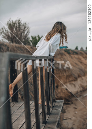 unrecognizable woman in white shirt standing barefoot on wooden platform with one foot raised, natural cliff landscape, back view, solitude, personal reflection, nature retreat 126562500