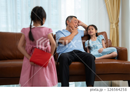 Little girl hiding present box behind back while making surprise elderly grandparents, Cheerful elderly man smiling and gesticulating while receives a surprise gift from granddaughter for celebration Little girl hiding present box behind back while making surprise elderly grandparents, Cheerful elderly man smiling and gesticulating while receives a surprise gift from granddaughter for celebration 126564258