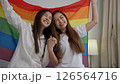 Portraits of happy Asian lesbian couple sitting with rainbow flag sitting bed beside the window. Cheerful two young adult women living together with relationship concept. LGBTQ Lifestyle Pride Month 126564716