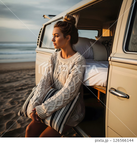 A female, about 25 years old, sits by an open van on a beach at sunset, wearing a sweater and holding a striped pillow 126566144
