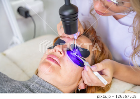 Close up view of eyelash extension procedure using ultraviolet lamp in modern beauty salon. Innovative lash enhancement technology with precise application technique. 126567319