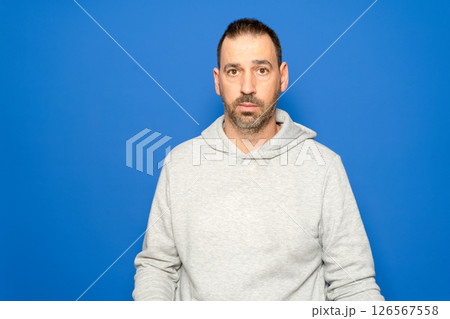 Hispanic man with a beard in his 40s wearing a gray sweatshirt looks straight ahead with a serious and worried expression, isolated on a blue studio background. 126567558