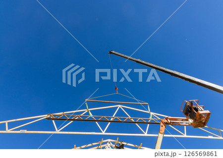 Crane raises large trusses framework component high into air in bustling construction zone. Crane raises large trusses framework component high into air in bustling construction zone. 126569861