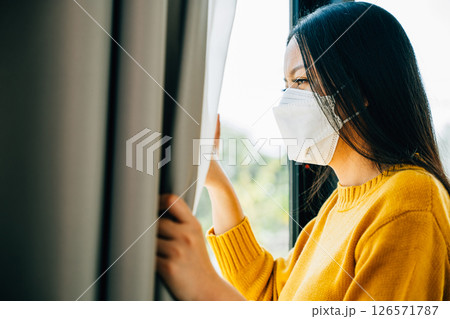 A young Asian woman in a medical mask looks through the window feeling upset and distressed due to COVID-19. Reflecting fear isolation and pandemic-related anxiety. A young Asian woman in a medical mask looks through the window feeling upset and distressed due to COVID-19. Reflecting fear isolation and pandemic-related anxiety. 126571787