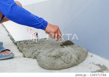 A worker hand uses a trowel to smooth gray mortar, preparing a level surface for floor tiles. Precision and craftsmanship in tiling work are on display. A worker hand uses a trowel to smooth gray mortar, preparing a level surface for floor tiles. Precision and craftsmanship in tiling work are on display. 126571788