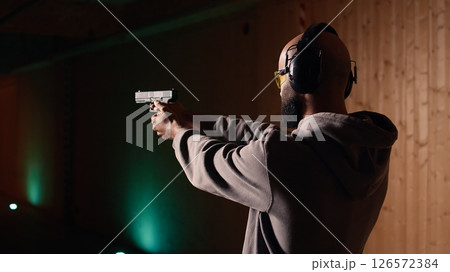 Shooting range customer shooting targets with pistol, learning to operate handgun in close quarters. Man at indoor shooting ground firing lethal fire weapon, wearing protection equipment, camera A Shooting range customer shooting targets with pistol, learning to operate handgun in close quarters. Man at indoor shooting ground firing lethal fire weapon, wearing protection equipment, camera A 126572384