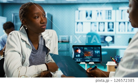 Radiologist delivering bad news about a tumor during a medical consultation, using CT scan data and microscopic illustrations to discuss surgery and cancer treatment advancements. Camera B. 126572385