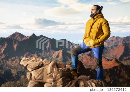 Adventure seeker gazes at the valley and mountain peaks ahead, feeling the freedom of the open wilderness. Young explorer fully equipped enjoying high altitude outdoor exploration. 126572412