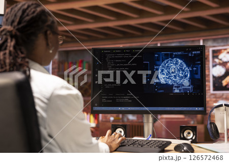 Teleworking system administrator using computer for artificial intelligence computing through self learning algorithms. African american remote worker works with AI deep neural networks on desktop PC 126572468