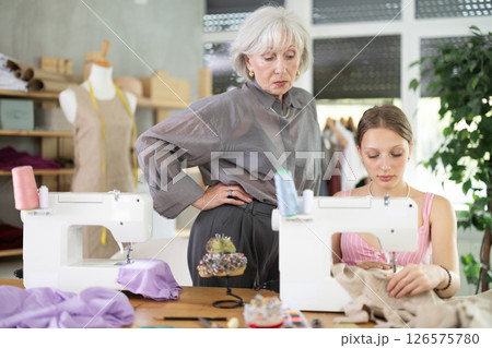 Elderly woman seamstress teaches young woman to sew Elderly woman seamstress teaches young woman to sew 126575780