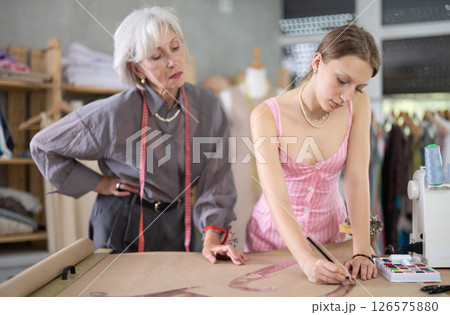 Elderly woman seamstress teaches young woman to draw pattern on paper 126575880