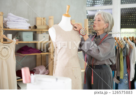 Mature female dressmaker working with mannequin in sewing workshop Mature female dressmaker working with mannequin in sewing workshop 126575883