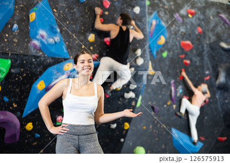 Fit young woman in sportswear smiling at camera while standing against artificial training wall in adventure park or rock climbing gym Fit young woman in sportswear smiling at camera while standing against artificial training wall in adventure park or rock climbing gym 126575913