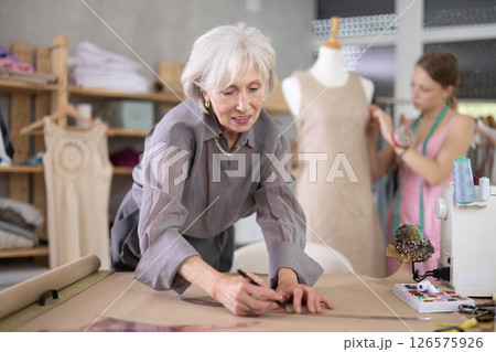 Mature tailor draws of a fashion design against the background of a assistant Mature tailor draws of a fashion design against the background of a assistant 126575926