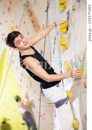 young positive man, insured by trainer, moving up artificial climbing wall indoors 126575965