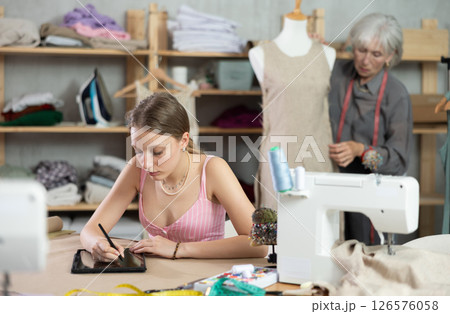Young woman draws pattern on tablet female assistant sews on mannequin Young woman draws pattern on tablet female assistant sews on mannequin 126576058