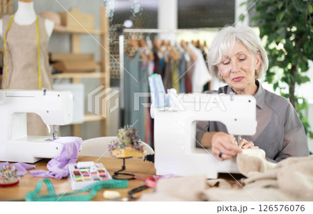 Mature female dressmaker working with sewing-machine in tailor's workshop Mature female dressmaker working with sewing-machine in tailor's workshop 126576076