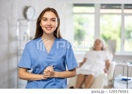 Smiling senior woman doctor in uniform with woman patient lying behind in clinic. Smiling senior woman doctor in uniform with woman patient lying behind in clinic. 126576081