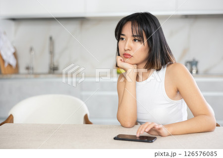 Sad young Asian girl sitting at the kitchen table with smartphone in hand 126576085