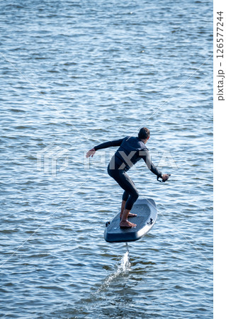 Surfer balances on electric hydrofoil board, gliding above rippling water with arms outstretched Surfer balances on electric hydrofoil board, gliding above rippling water with arms outstretched 126577244