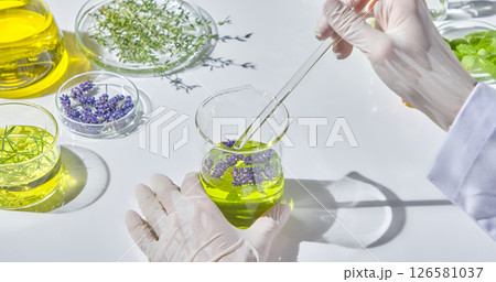 A researcher's hand is stirring a bright green natural oil mixture in a glass beaker with a glass stirring rod, surrounded by fresh and dried herbal ingredients. A researcher's hand is stirring a bright green natural oil mixture in a glass beaker with a glass stirring rod, surrounded by fresh and dried herbal ingredients. 126581037