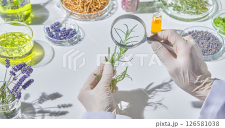 Holding a small glass vial of green botanical oil and a sprig of rosemary, with scientific tools and dried herbs in background.Rosemary, lavender, tea tree... are all herbs with many uses. Holding a small glass vial of green botanical oil and a sprig of rosemary, with scientific tools and dried herbs in background.Rosemary, lavender, tea tree... are all herbs with many uses. 126581038