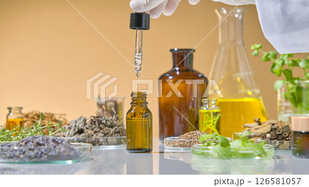 Close-up of essential oil being extracted from amber glass bottle with dropper in natural science research setup. White lab table with herbs displayed around it Close-up of essential oil being extracted from amber glass bottle with dropper in natural science research setup. White lab table with herbs displayed around it 126581057