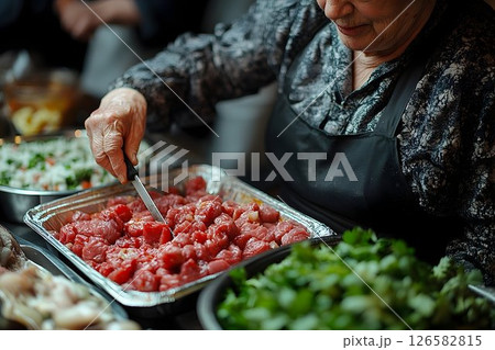 Elderly woman carefully cuts raw meat in a foil tray, preparing 126582815