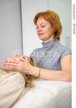 Female practitioner performs facial lifting technique on a young woman lying on a couch. Concept of energy healing, face therapy and alternative wellness practice from Access Bars. Female practitioner performs facial lifting technique on a young woman lying on a couch. Concept of energy healing, face therapy and alternative wellness practice from Access Bars. 126583561