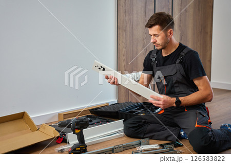 Man in overalls assembles furniture, examining white panel and surrounded by tools. Handyman in work clothes preparing parts pf cabinet to assembly. DIY furniture project Man in overalls assembles furniture, examining white panel and surrounded by tools. Handyman in work clothes preparing parts pf cabinet to assembly. DIY furniture project 126583822