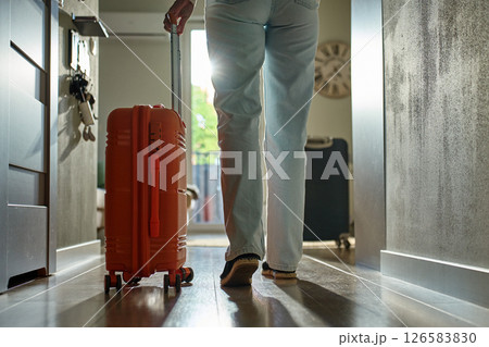 Low-angle view of traveler walking towards door with rolling luggage. Person walks across hallway with orange suitcase. Concept of departure and leaving home for vacation 126583830