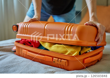 Woman trying to close suitcase at home. Female press down overfilled orange suitcase while packing clothes. Person preparing for departure Woman trying to close suitcase at home. Female press down overfilled orange suitcase while packing clothes. Person preparing for departure 126583848